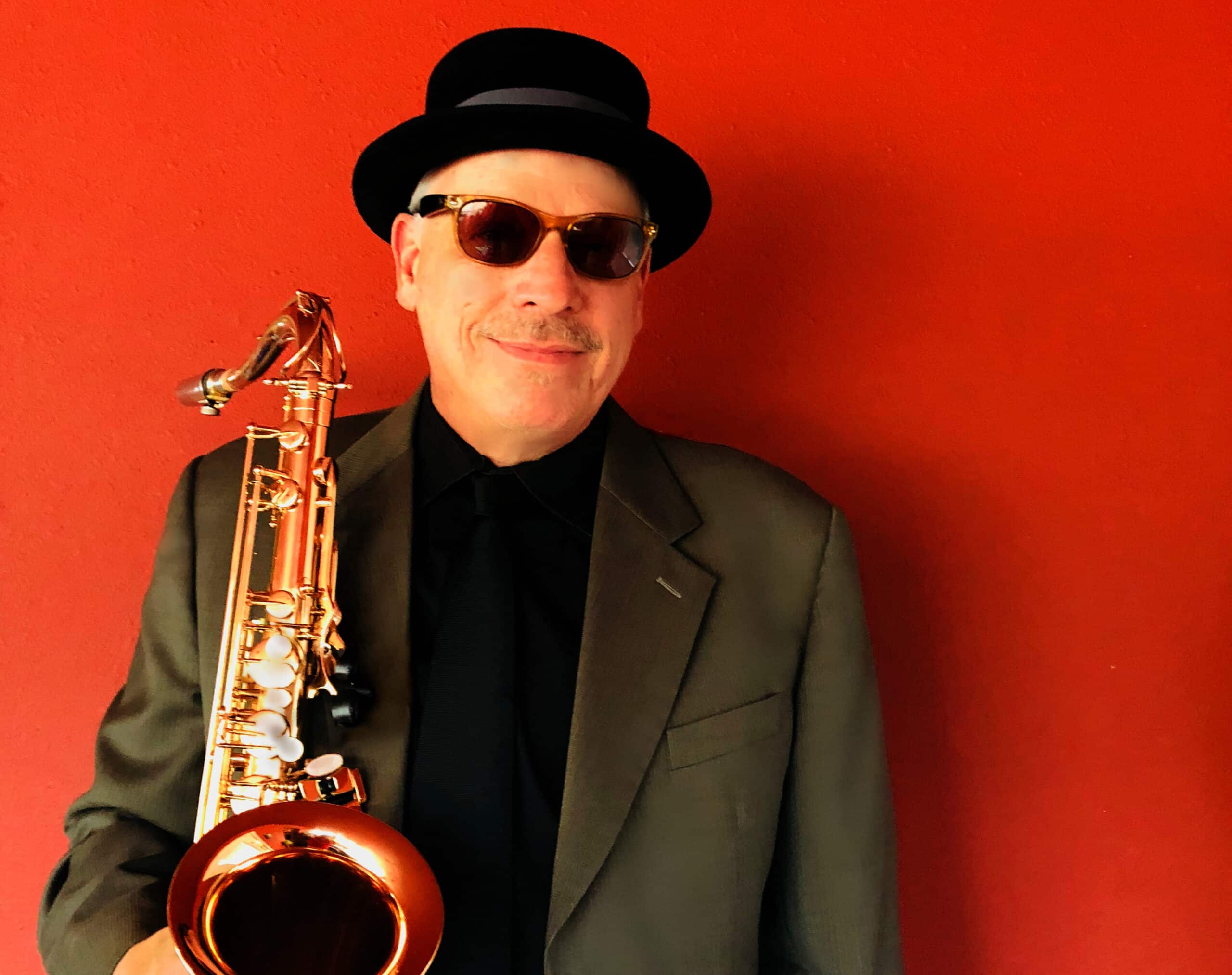 Doug Lawrence stands in front of a red background with his saxophone, wearing a suit jacket, sunglasses and hat