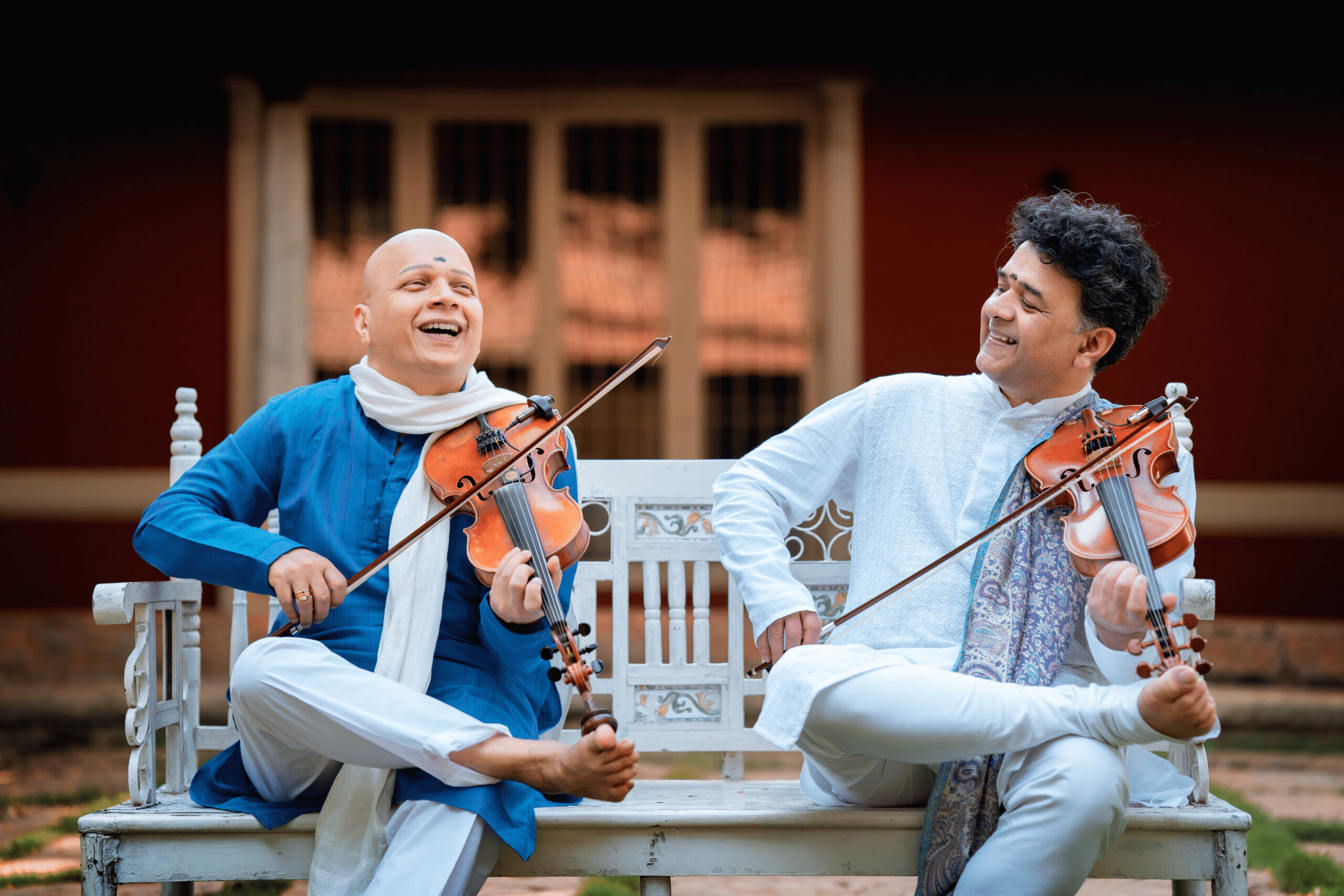 Ganesh and Kumaresh laugh to one another whilst playing their violins outdoors on a white bench