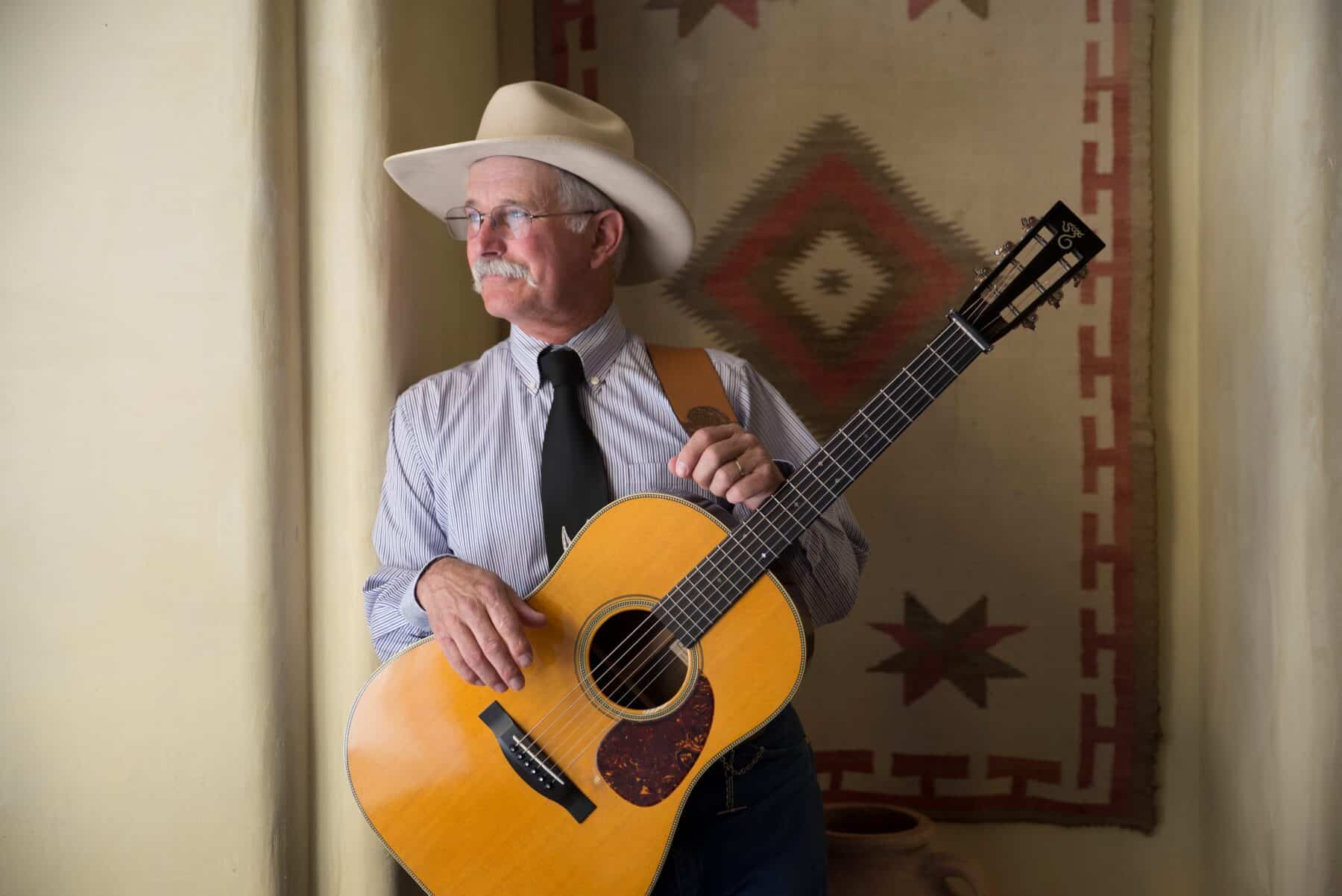 Dave Stamey is wearing a cowboy hat and holding a guitar as he leans against a wall in a Southwestern styled room