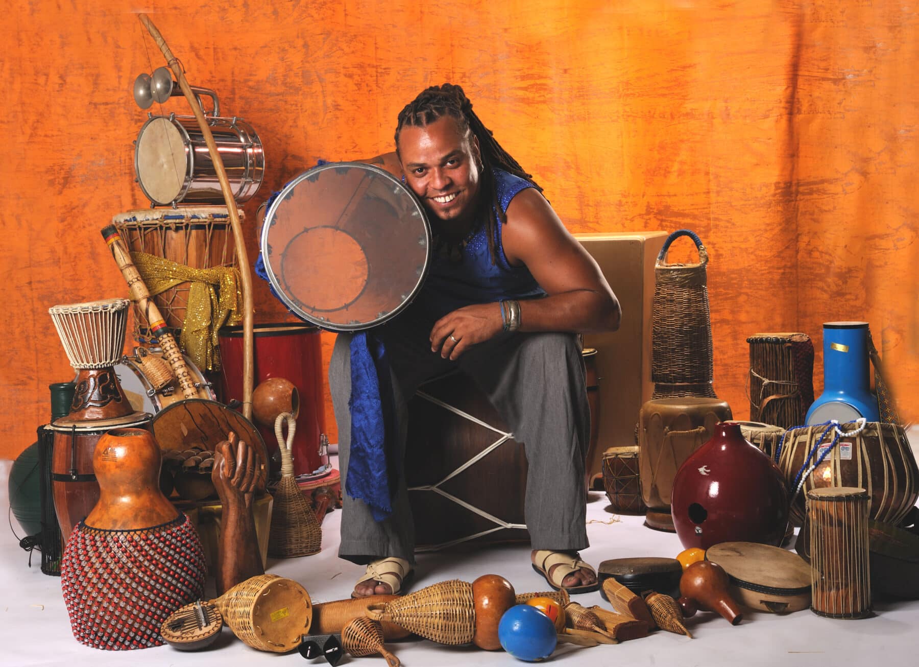 Dendê Macêdo is sitting on a drum laying on its side, holding another drum under his arm, and he is surrounded by various other instruments