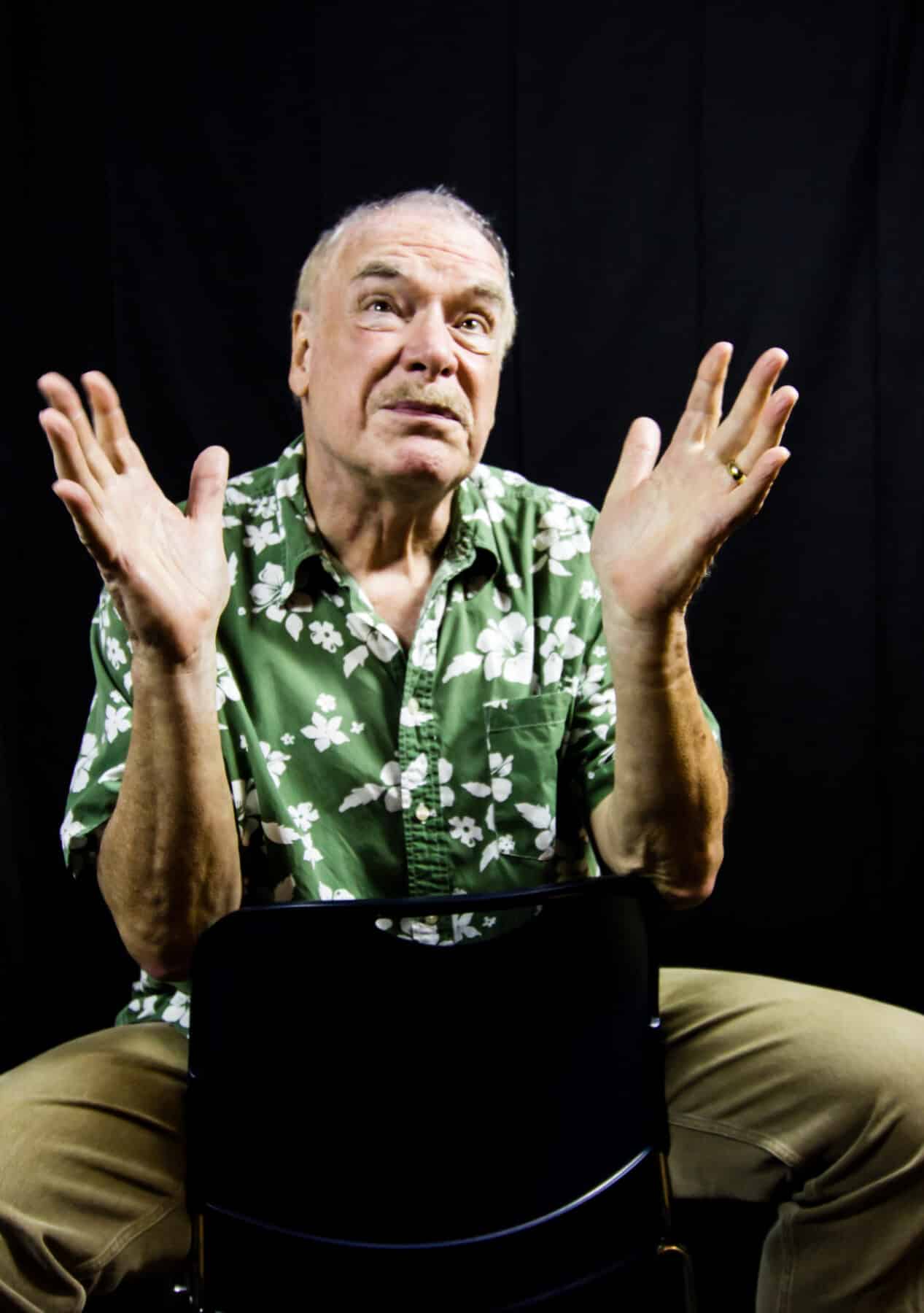 Jon Spelman is sitting on a chair, gesturing upwards with his hands, as he looks up and into the distance.
