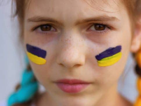 A young girl with face paint stripes resembling the Ukrainian flag stares into the camera. 
