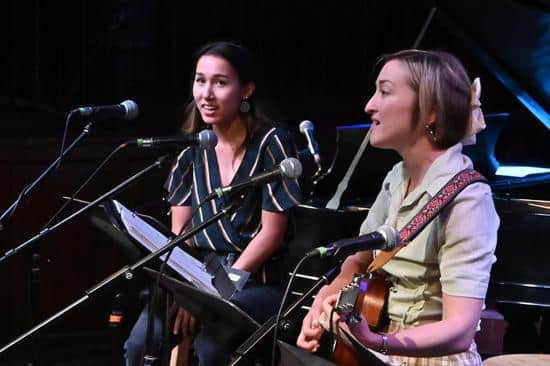 Two women sing on stage, while one also plays the guitar. They are sitting behind music stands and microphones.