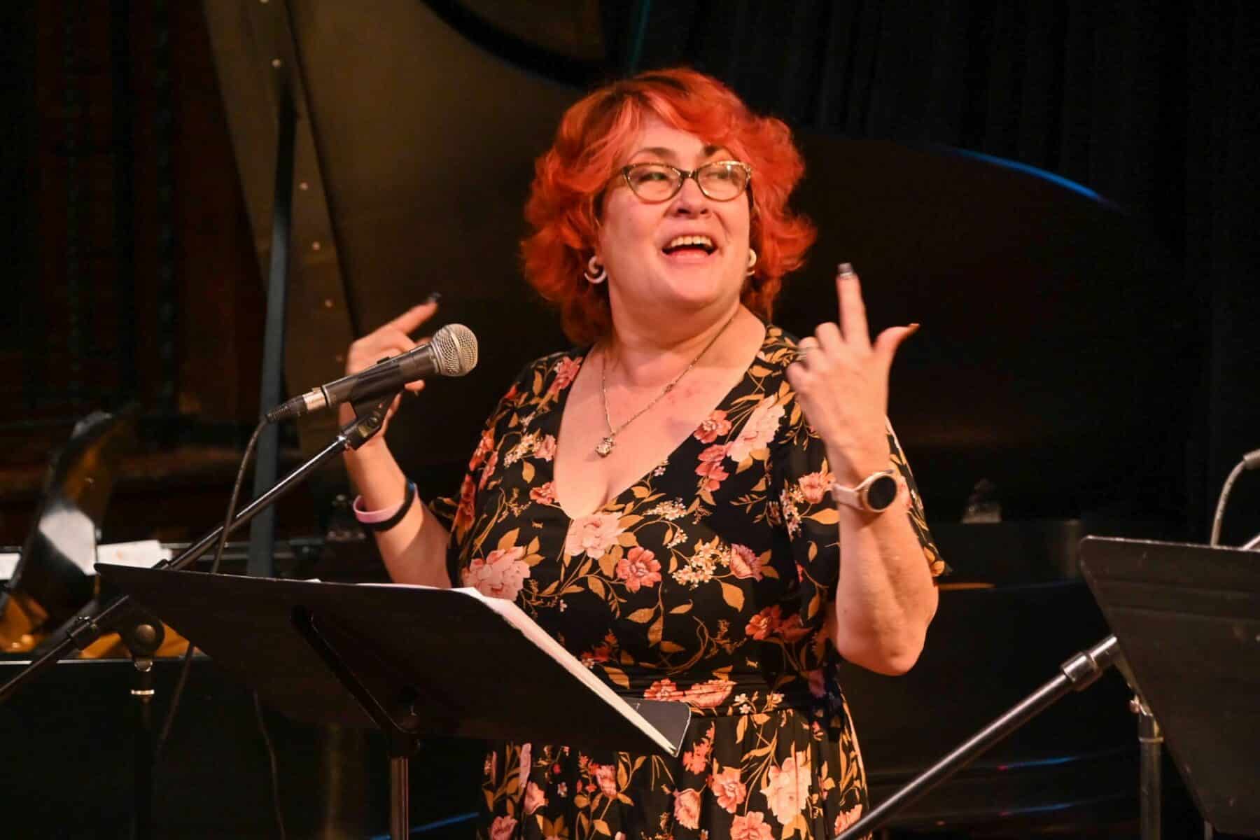 A woman with short red hair and glasses stands on stage, smiling as she gestures with her hands. She is wearing a floral dress and is in front of a microphone with a music stand.