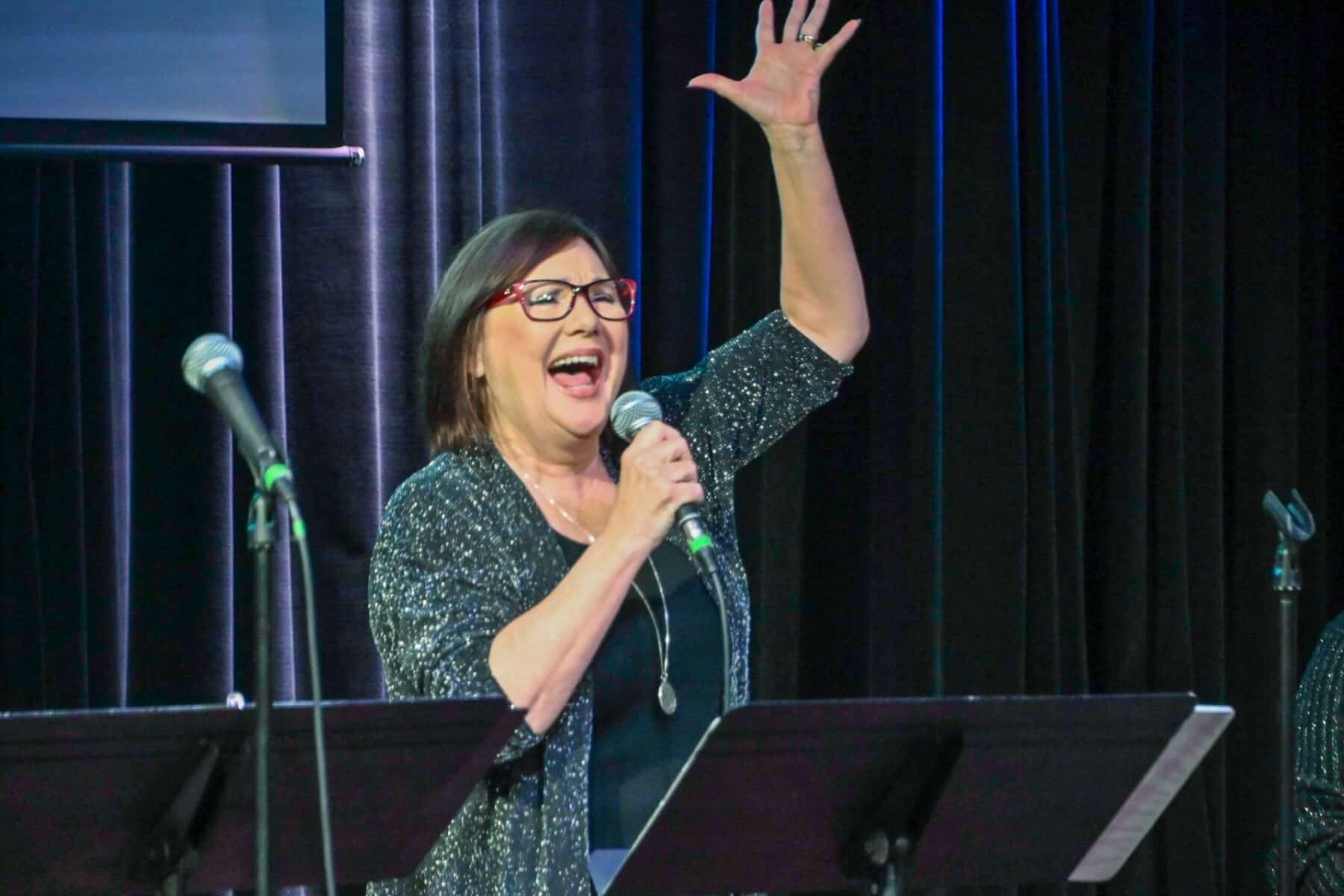 A woman sings on stage, holding her arms up. She is standing in front of a microphone with colorful lights in the background.