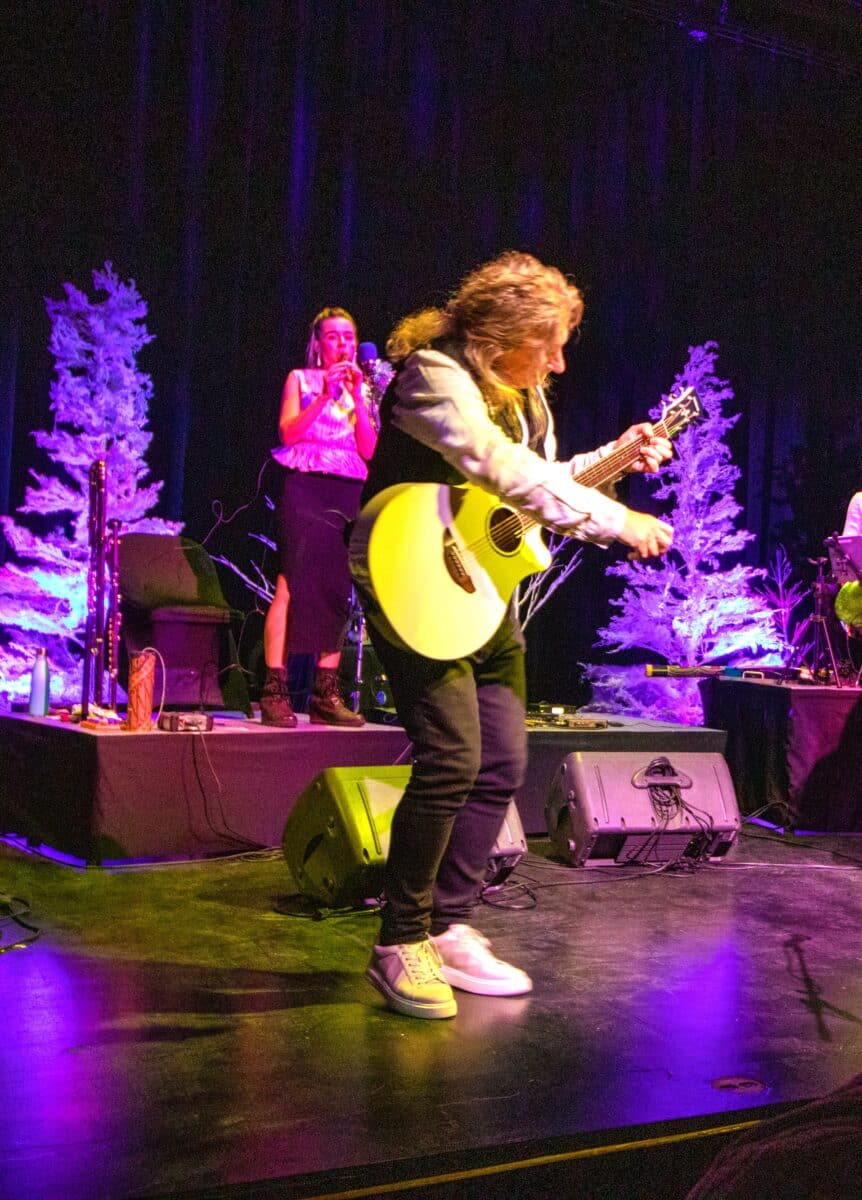 David Arkenstone plays guitar an acoustic guitar amid winter stage decor including white pine trees.