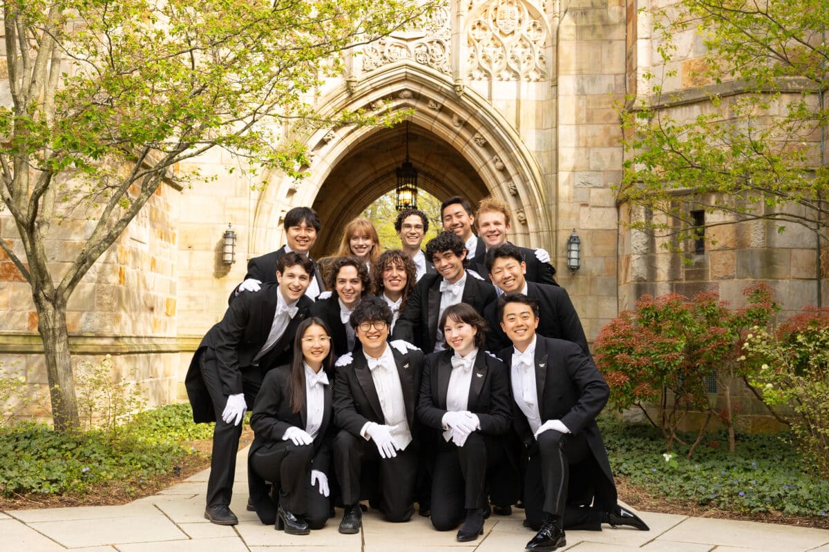 A capella group stands in front of a vaulted arch in gloves and tuxedos.