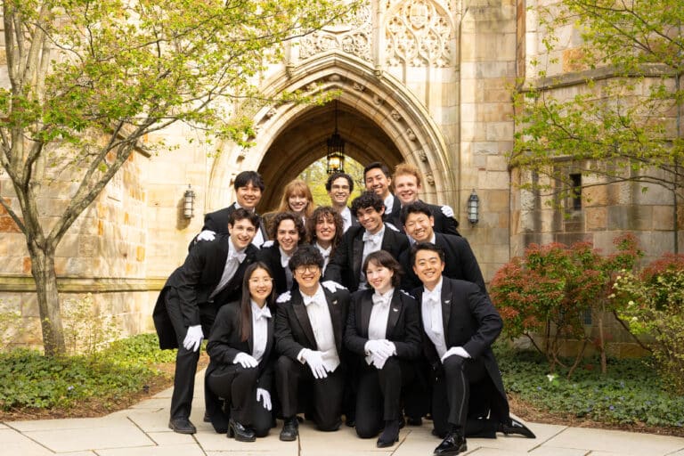 A capella group stands in front of a vaulted arch in gloves and tuxedos.