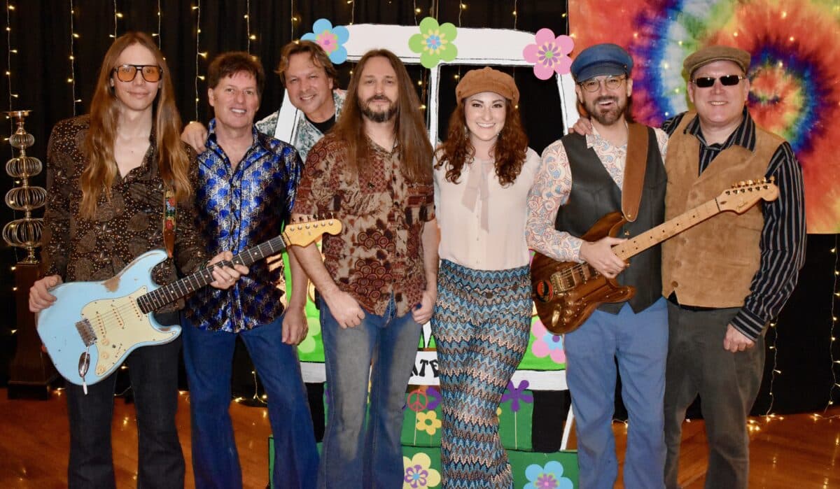Five band members dressed in '70s style clothing stand in front of a hippie van prop and rainbow tie-dye background