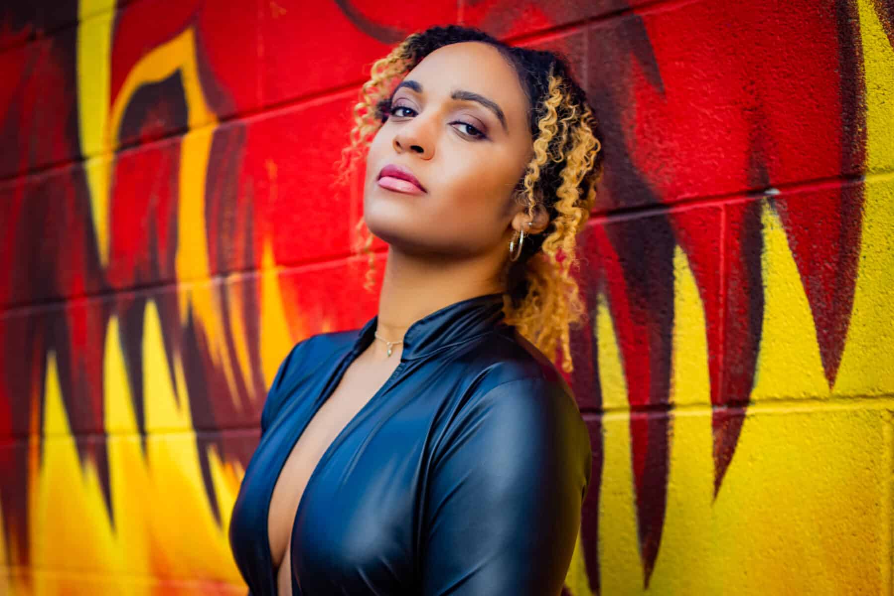 Portrait of a woman with curly highlighted hair standing against a bold red and yellow graffiti wall, wearing a black zip-front top and looking confidently toward the camera.