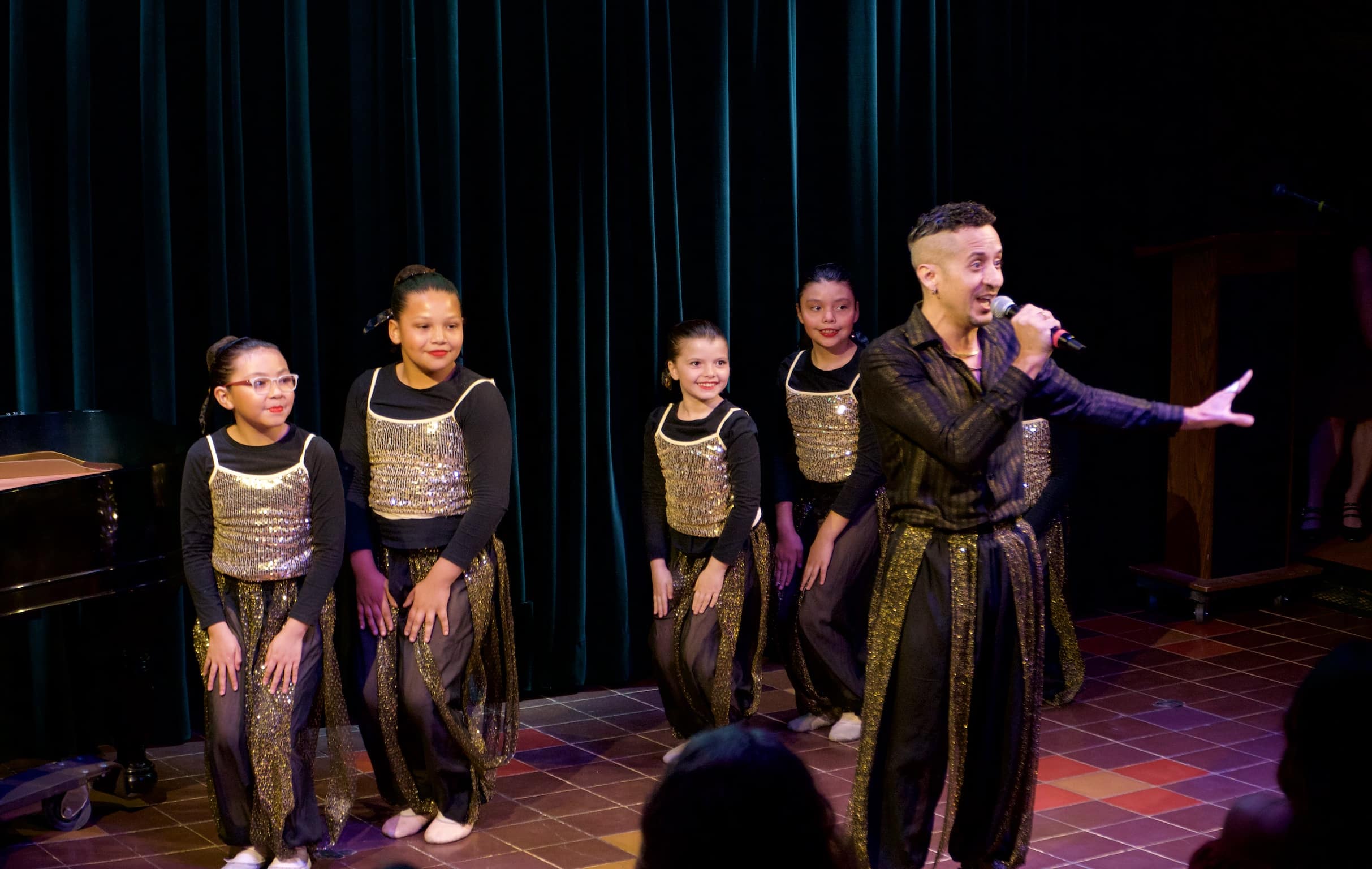 Male performer in a black-and-gold outfit sings into a microphone onstage, gesturing toward the audience as five young dancers in matching black costumes with gold sequined tops pose behind him in front of a dark curtain.