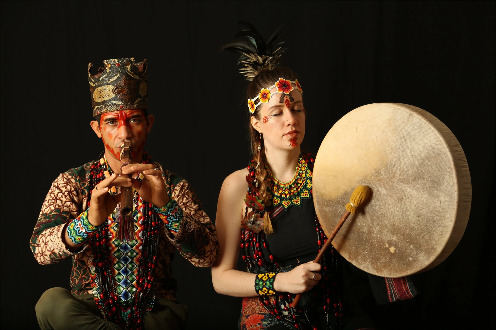 A man and a woman dressed in vibrant Amazonian-inspired traditional attire sit against a black background. The man plays a wooden flute and wears a patterned crown-like headpiece, face paint, and layered beaded necklaces. The woman, also wearing face paint, a feathered headpiece, and colorful beaded jewelry, holds and gently strikes a large hand drum with a padded mallet. Both appear focused and immersed in the music.