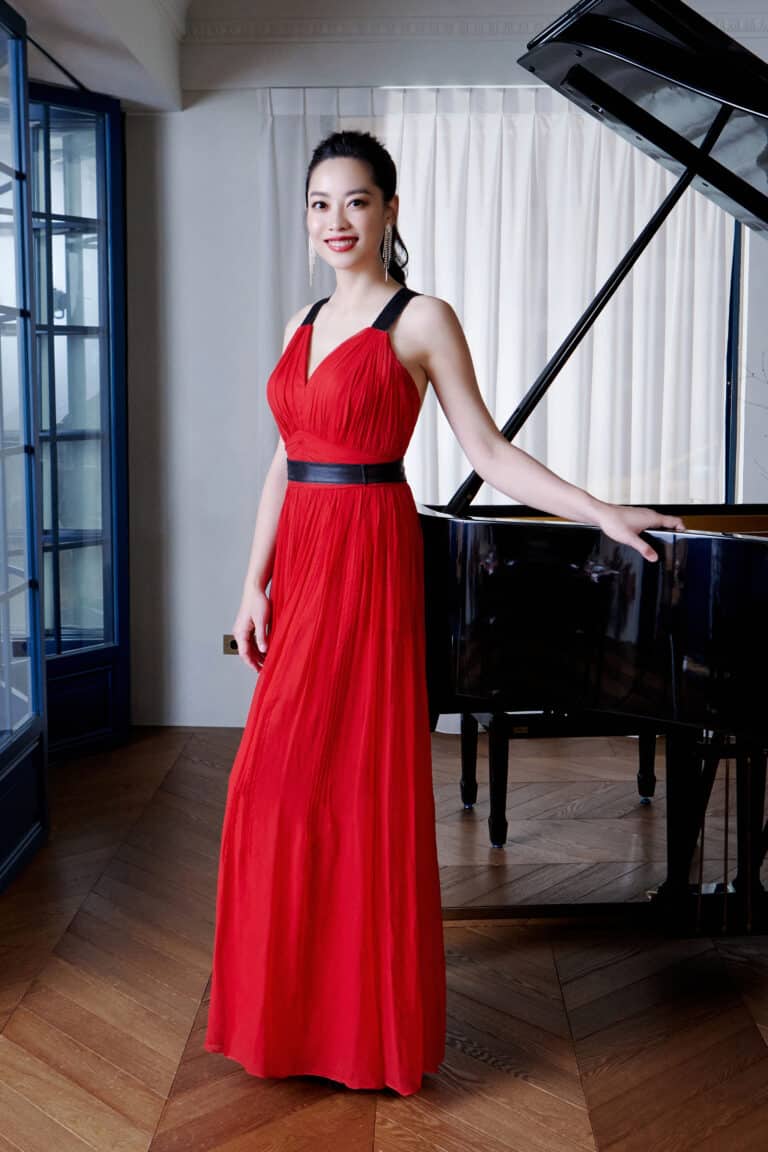 A woman in a red dress stands beside a piano, showcasing elegance and musicality in a stylish setting.