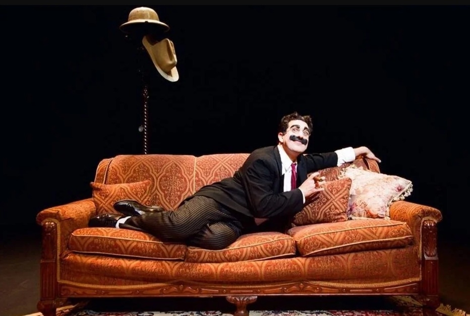 A dapper man in a suit and tie lounges on a couch, looking like he’s ready for a board meeting or a nap.