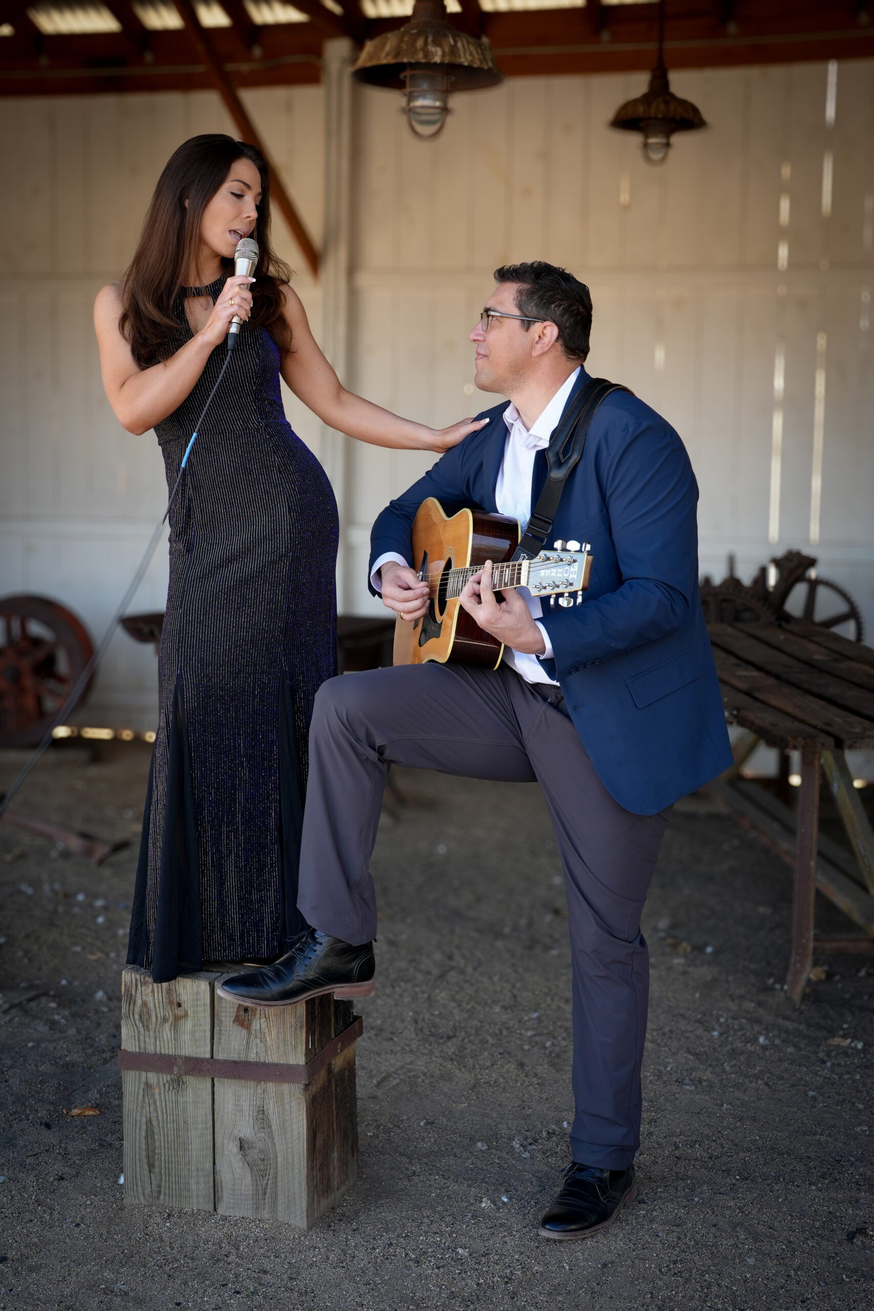 Emma stands on an elevated platform in a black gown, singing into a vintage microphone as James smiles below her wearing glasses, a blue blazer and grey pants whilst playing the guitar.