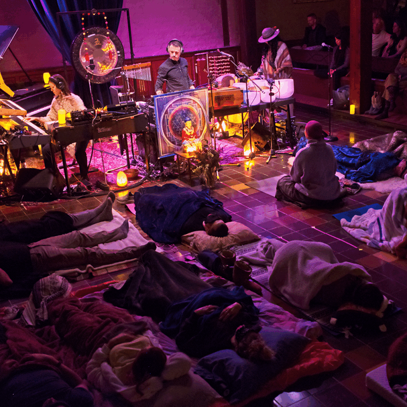 Spiritual Thing performs at Kerr with an expansive setup of synthesizers, grand piano, gong, crystal bowls, chimes and more. Salt lamps accent the stage area. The audience members are lying down on individual mats. One person is seated upright in a meditative pose.