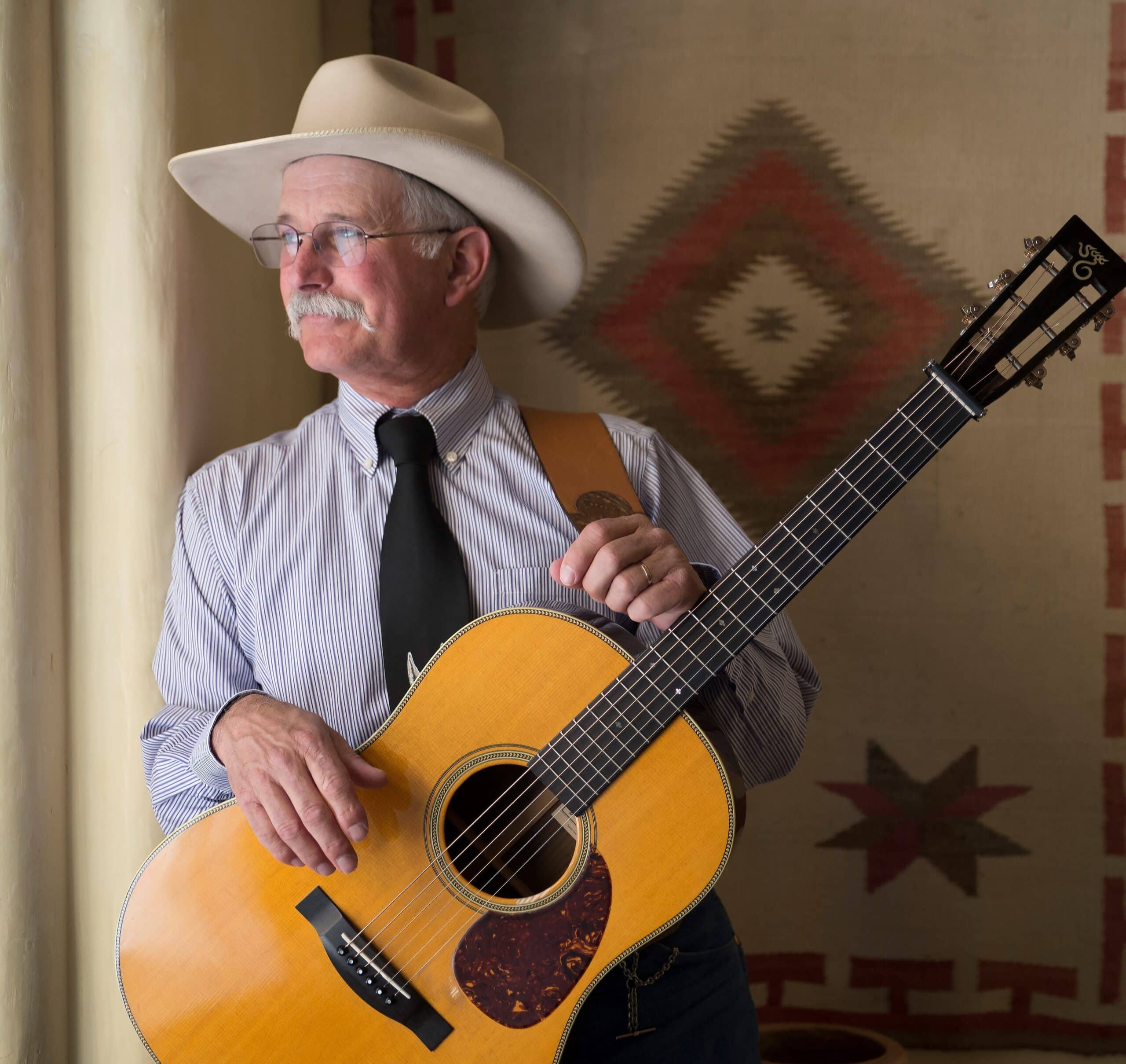 A man dressed in a crisp white shirt and cowboy hat holds a guitar and is looking confidently into the distance.