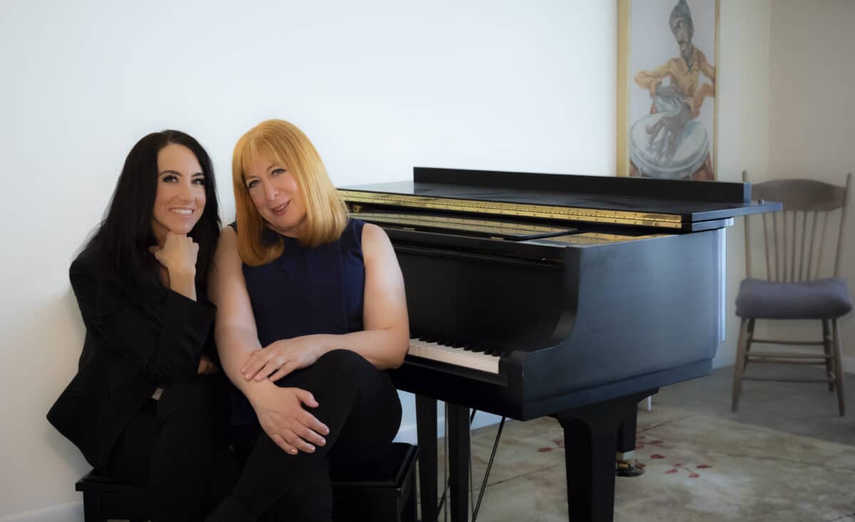 Two women sit beside a black piano, smiling in a light-filled room with artwork behind them.