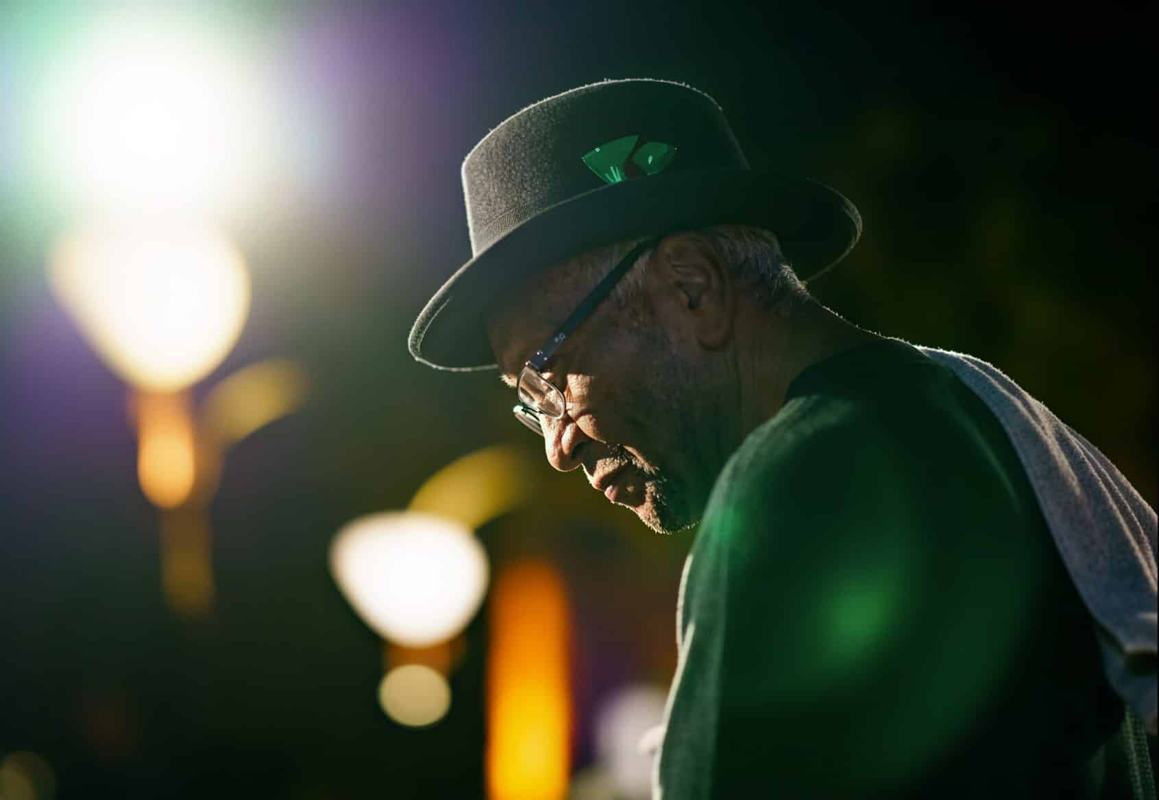 In a dimly lit space, a man wearing a hat and glasses gazes thoughtfully into the distance.