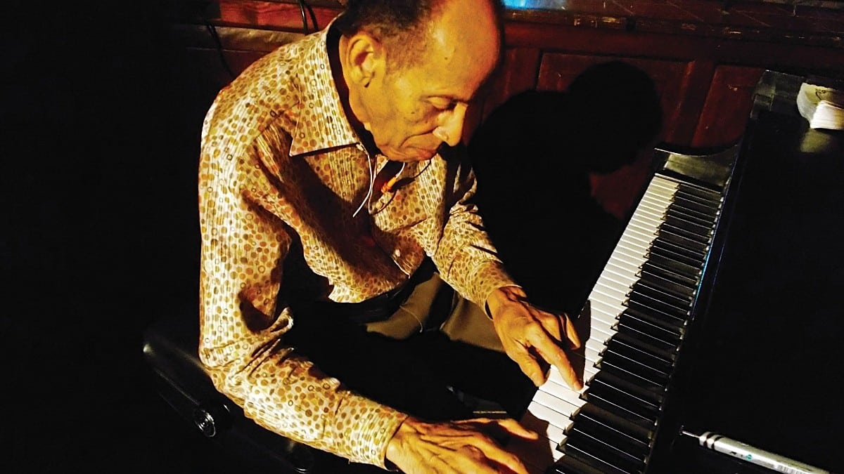 An older man in a patterned shirt plays the piano, focused on the keys in low light.