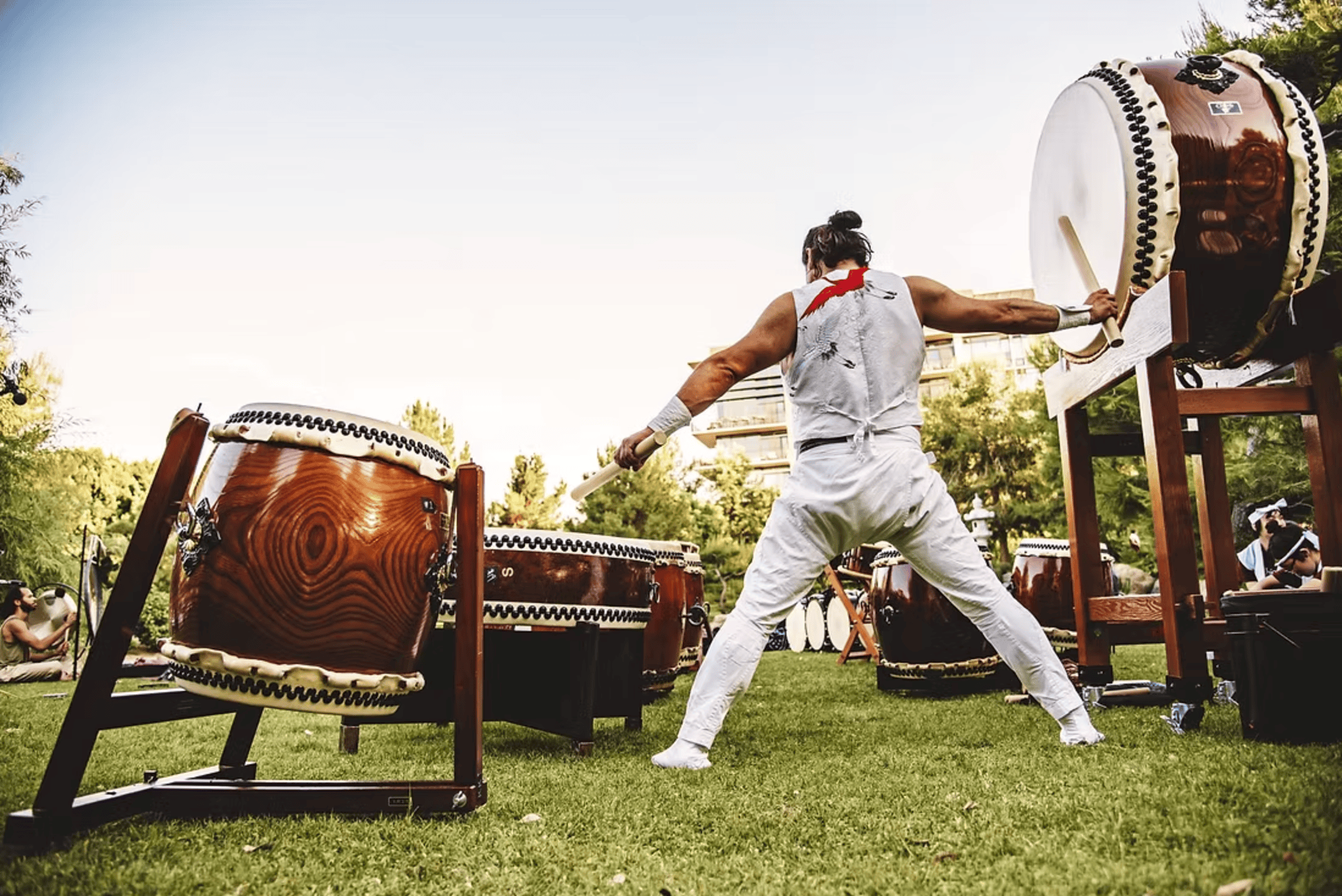 A man energetically playing drums while standing on grass, surrounded by a natural outdoor setting.