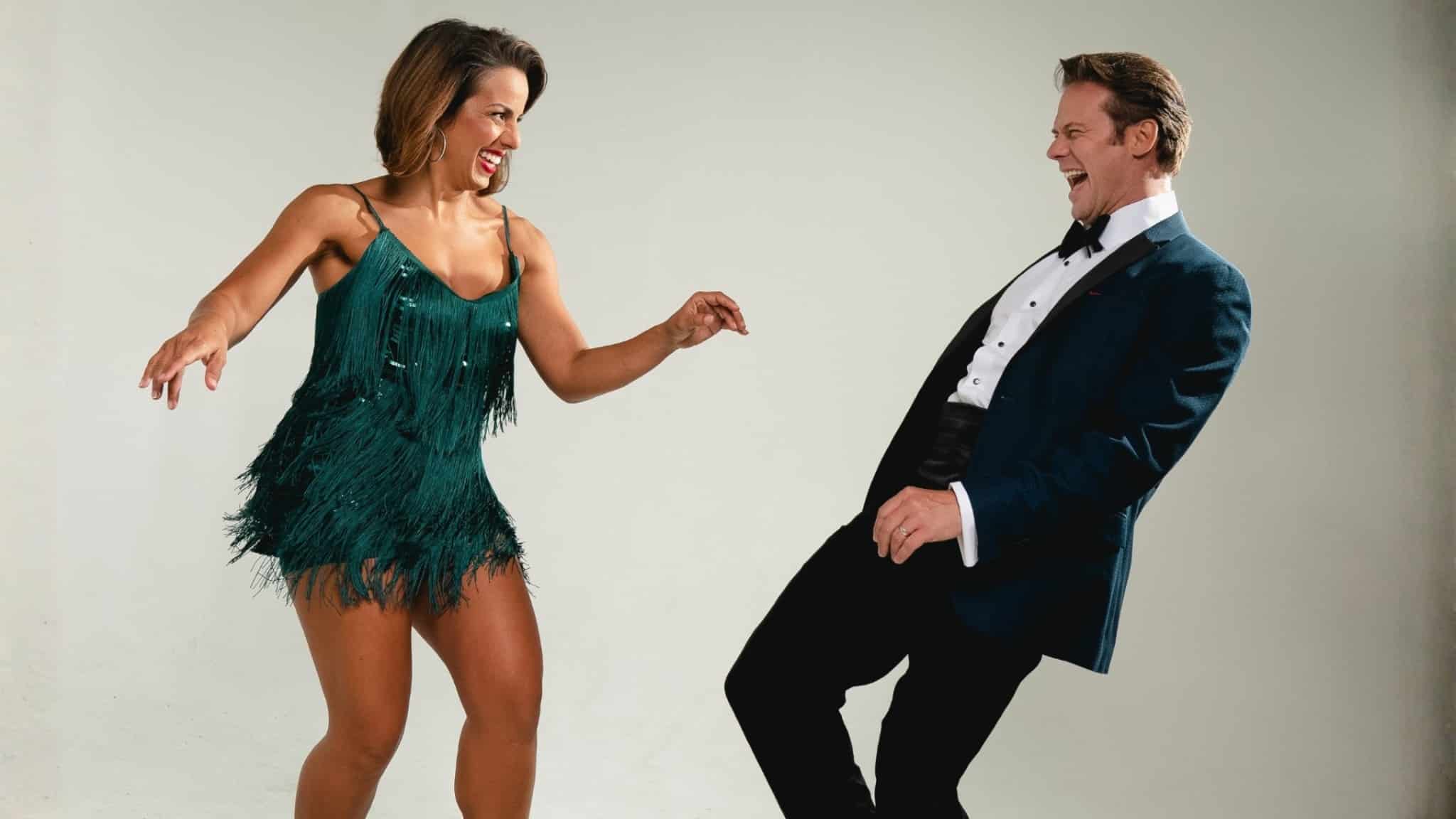 A woman in a green fringed dress and a man in a tuxedo smile and pose together as if dancing against a plain background.