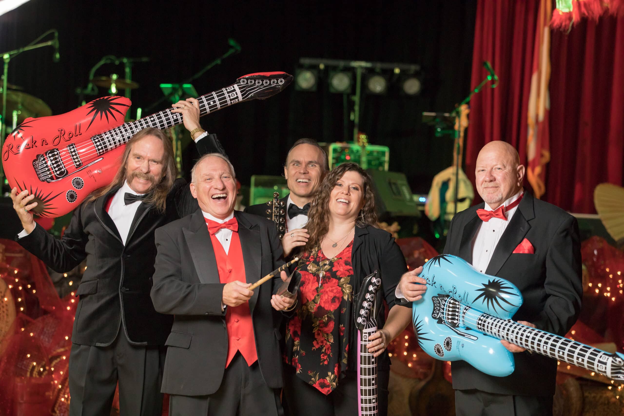 Five adults in formal attire pose and smile, holding inflatable guitars and drumsticks, with stage equipment and red curtains visible in the background.