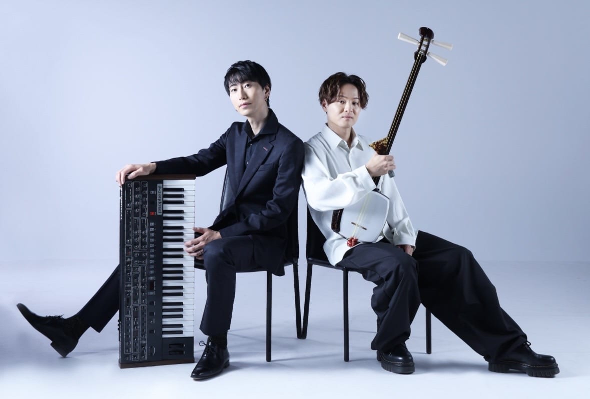 Two men sit back-to-back while holding instruments.