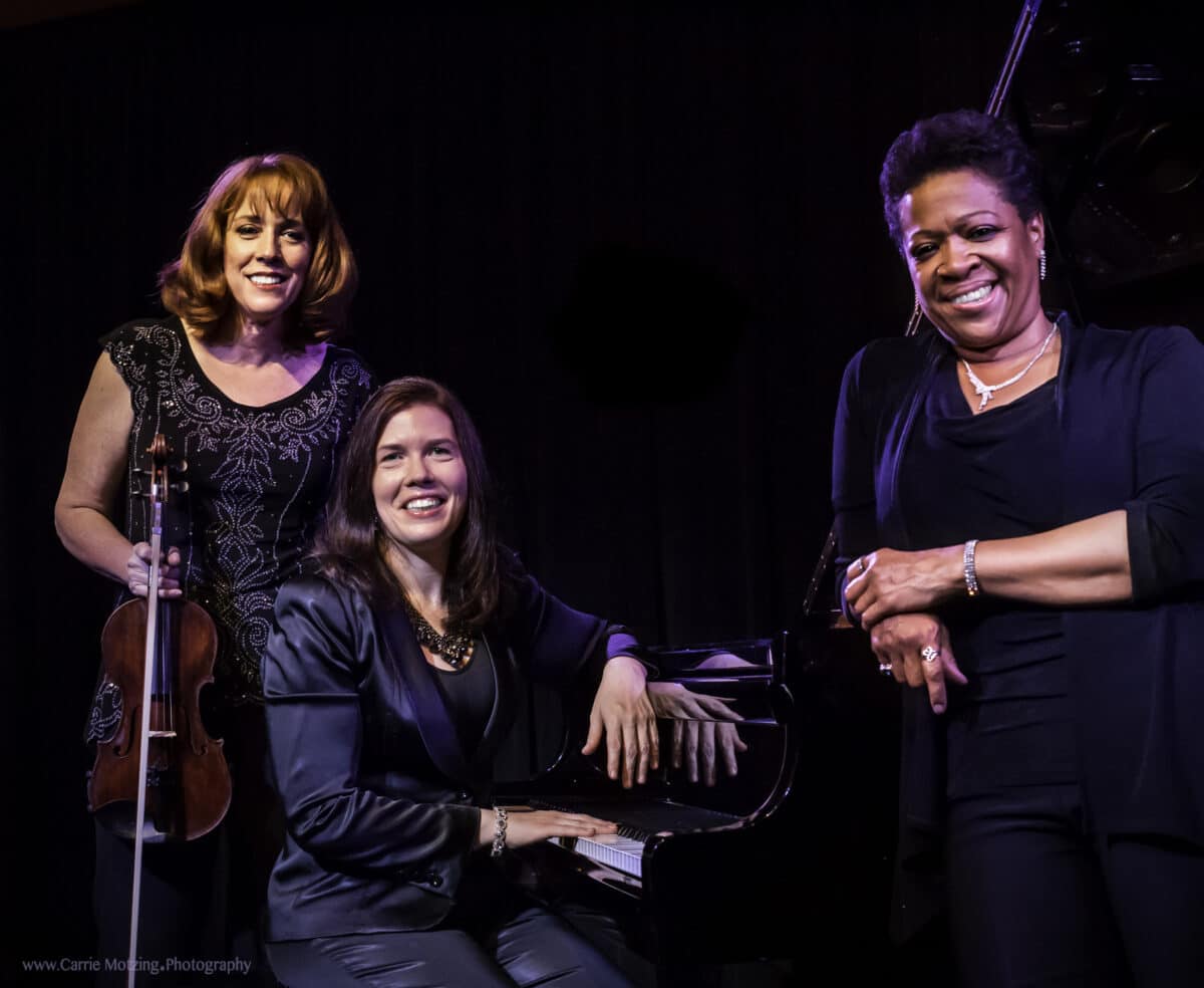 Three women smile at the camera while standing before a black background.