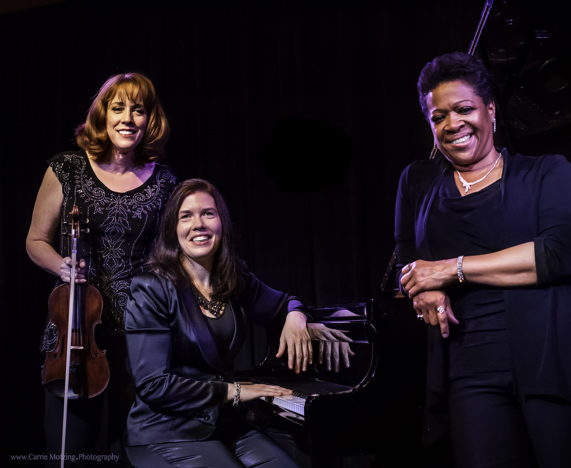 Three women smile at the camera while standing before a black background.