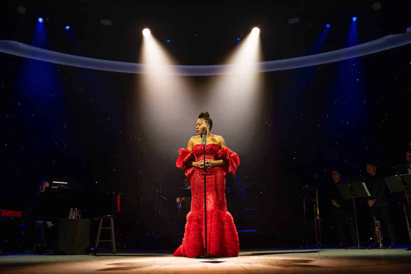 A woman wearing a red dress stands confidently on stage, engaging the audience with her presence.