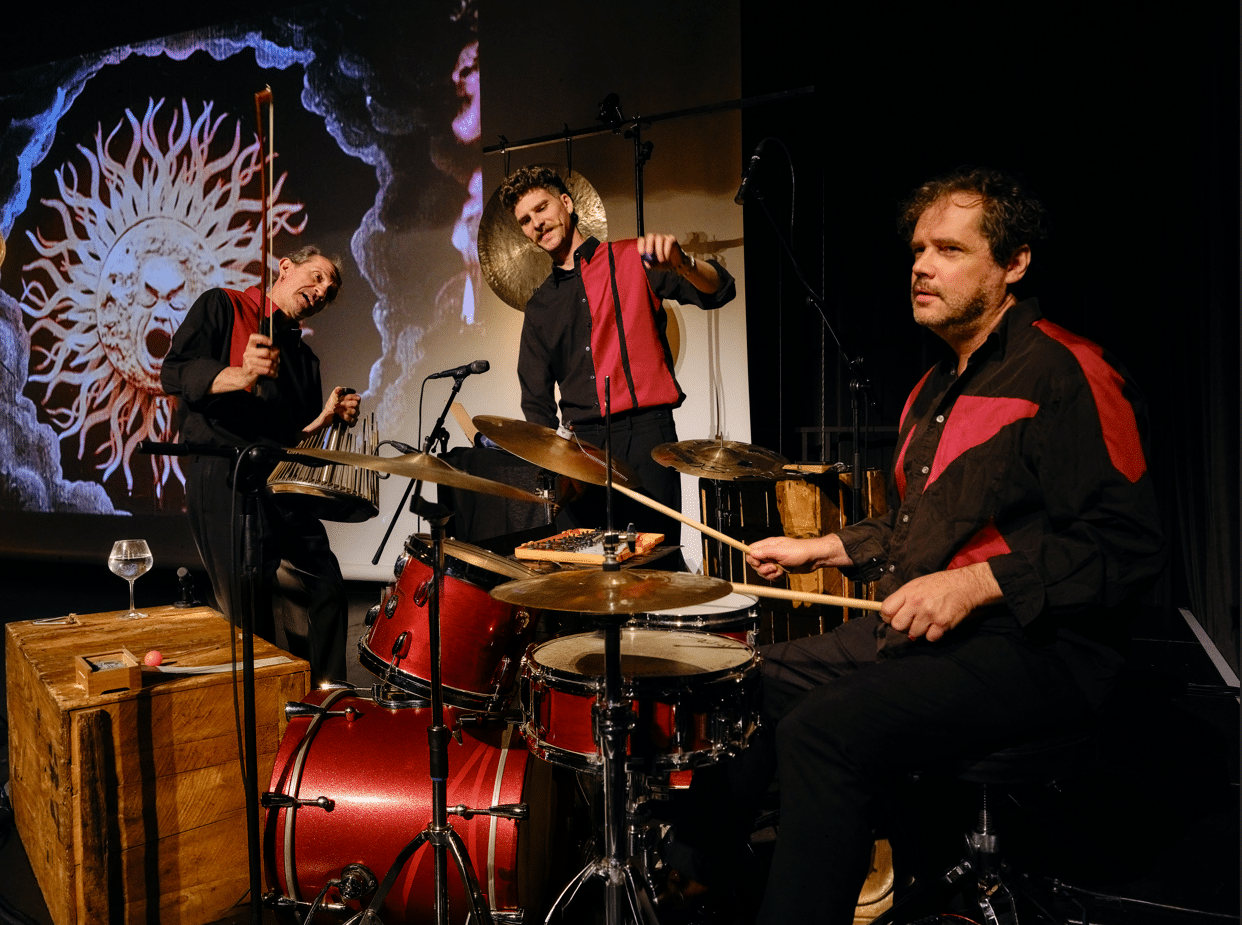 Three men performing on stage, playing drums, with a large projection screen displaying visuals behind them.