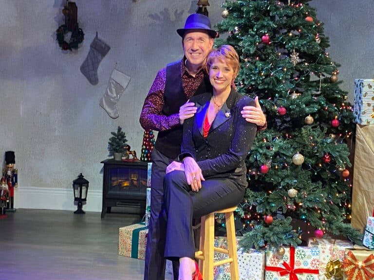 A man and woman stand close to each other while smiling at the camera in front of a Christmas tree.