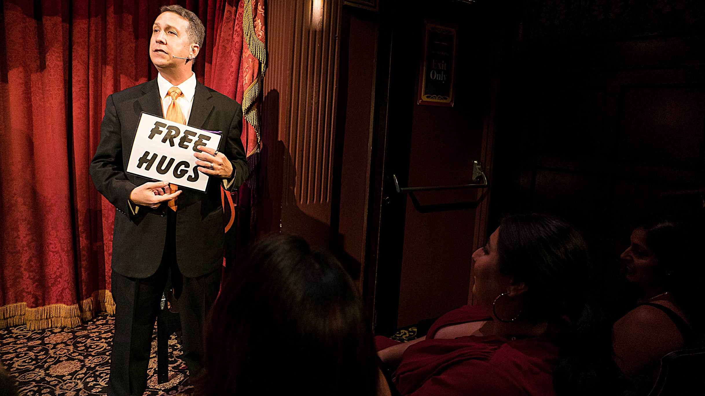 A man in a suit holding a sign that says "Free Hugs" stands on a stage in front of a bright red curtain.