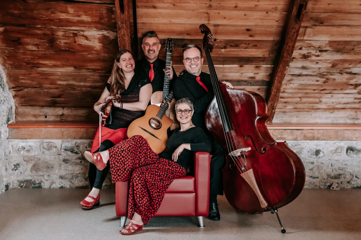 Four people sit together in a cabin-like setting while holding instruments such as a cello, guitar, and violin.