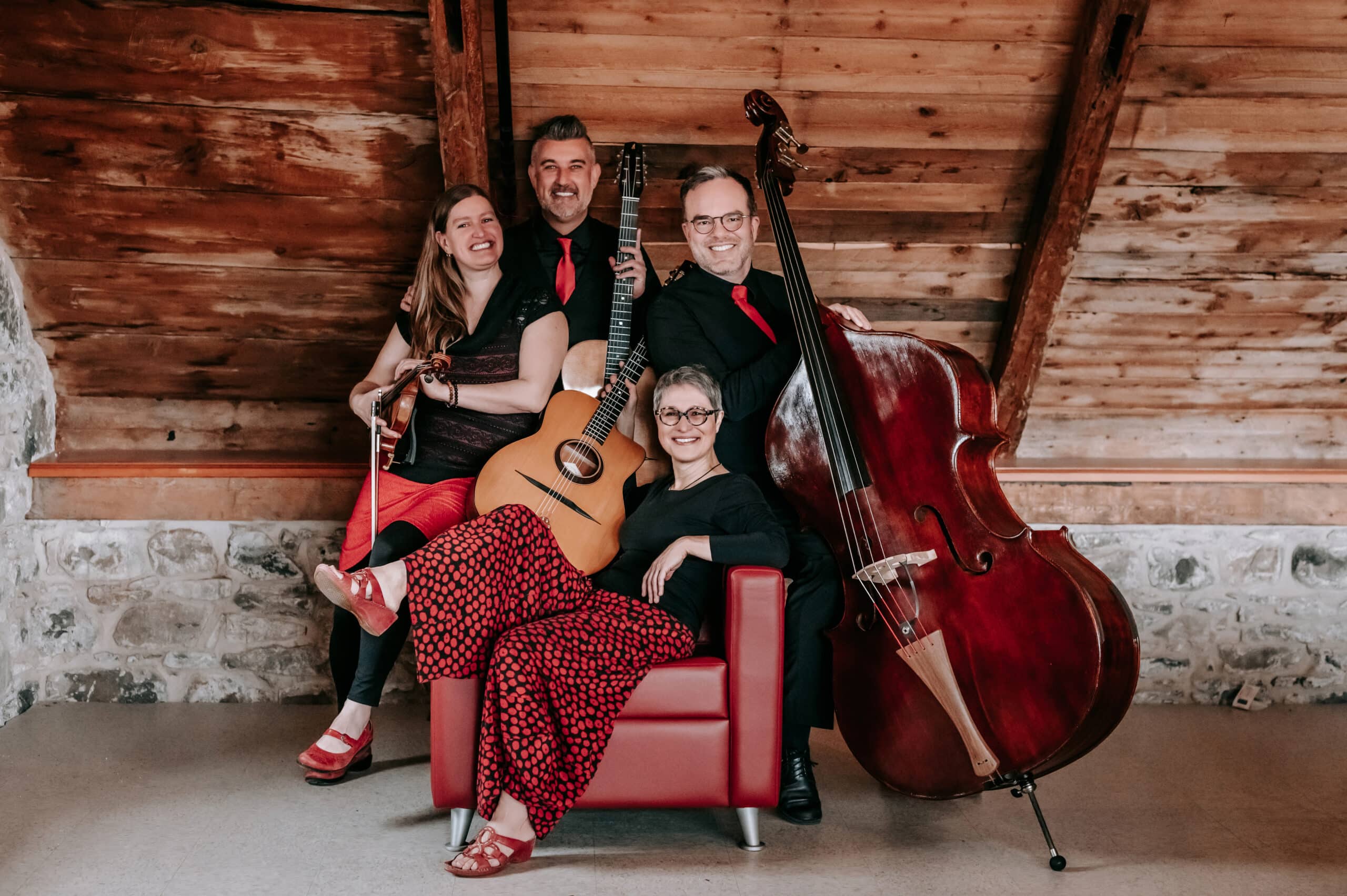 Four people sit together in a cabin-like setting while holding instruments such as a cello, guitar, and violin.